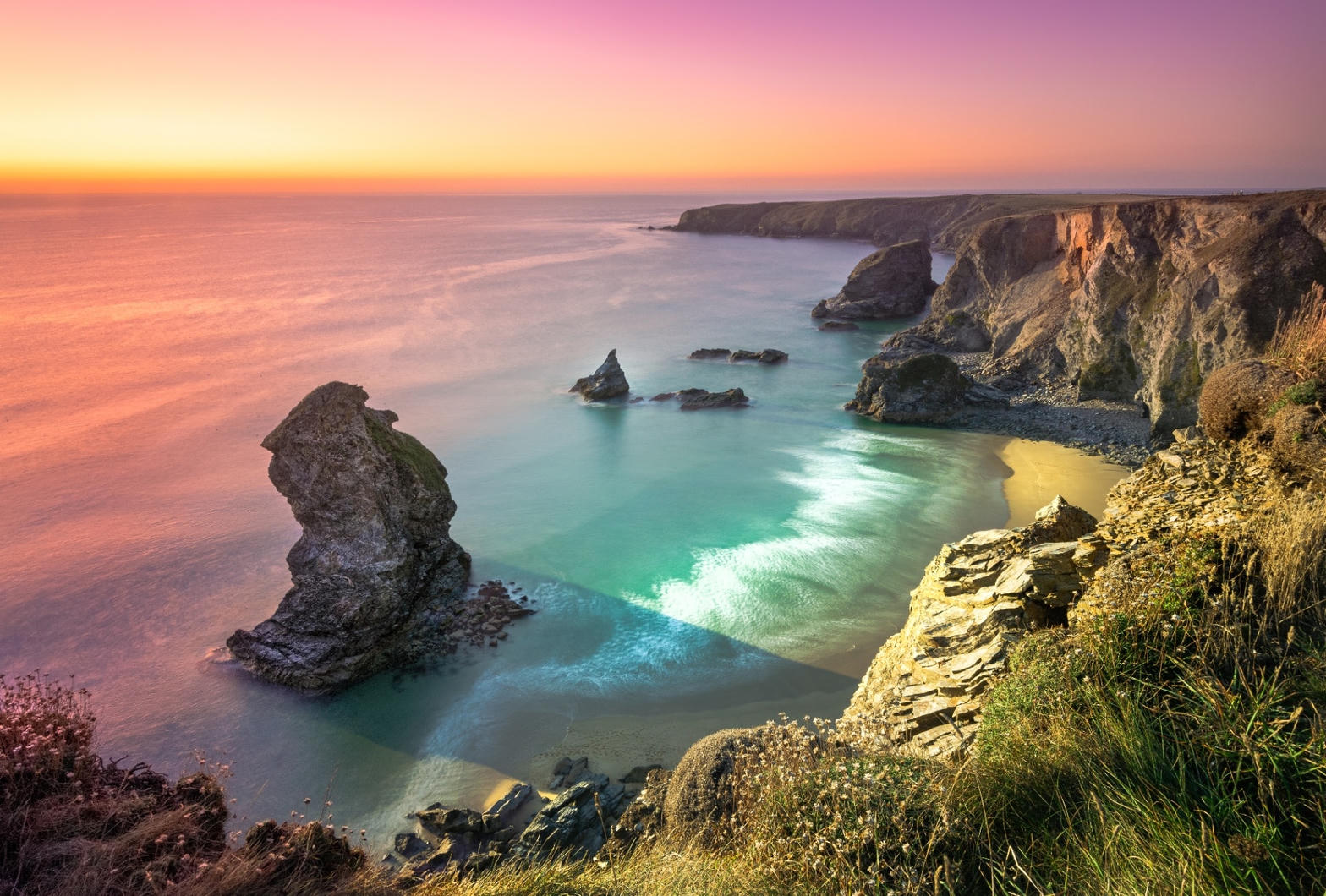 A Cornish cove at sunset, with a sea stack, turquoise water and cliff path.