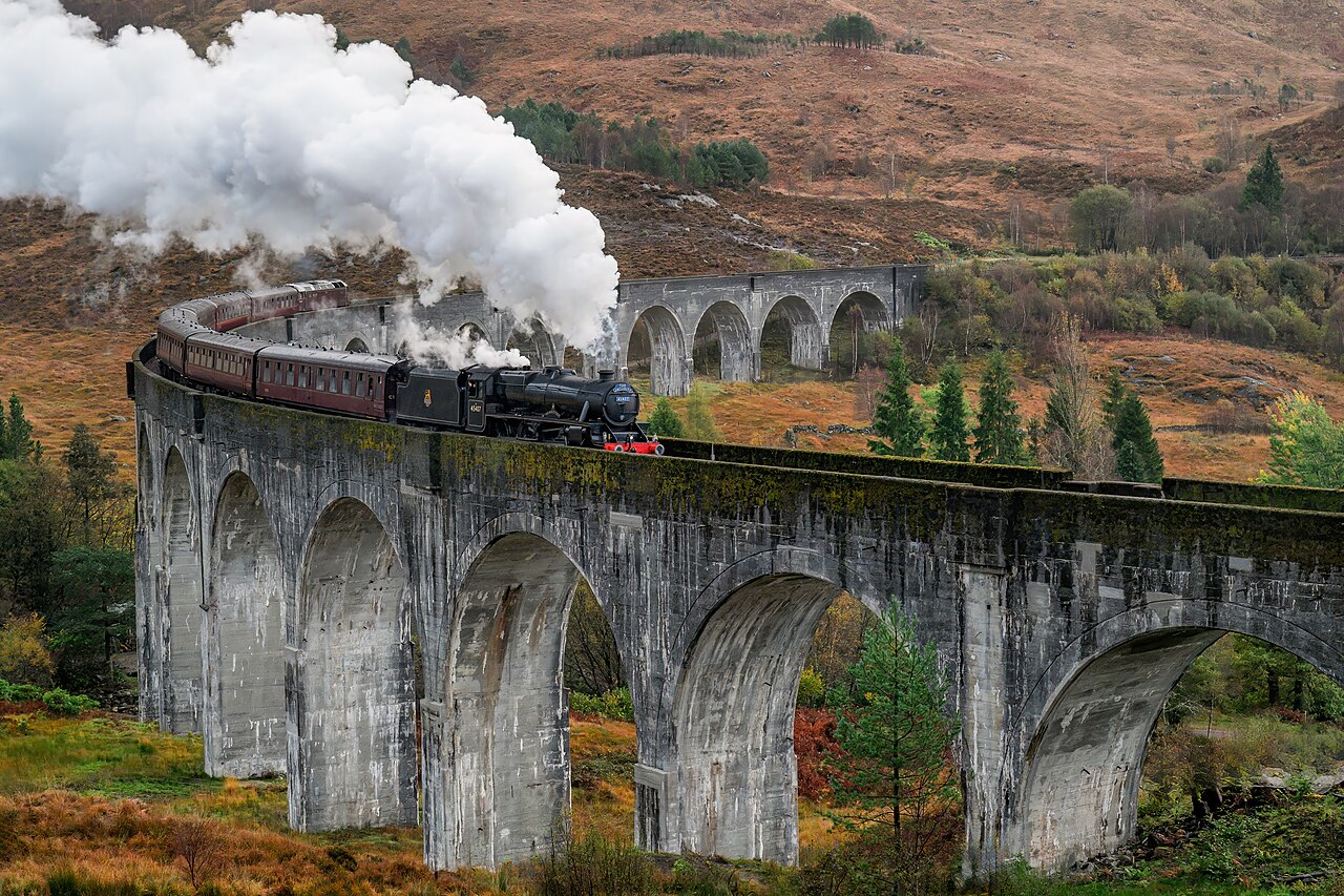 A steam train crossing the curved Glenfinnan Viaduct, with loch and hills beyond.